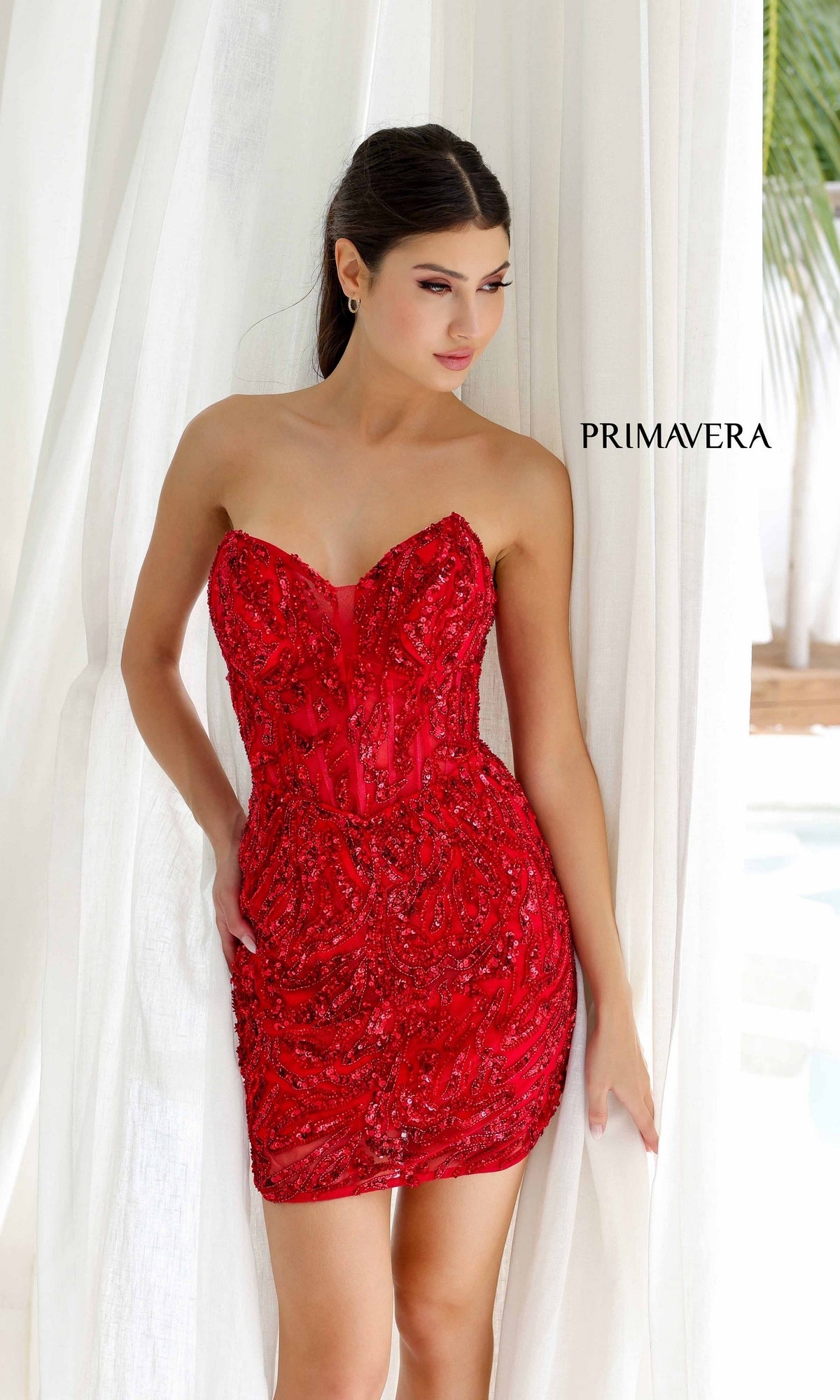 A woman stands before white curtains wearing Short Homecoming Dress 4222 by Primavera, a strapless homecoming dress featuring a corset bodice and red beading;